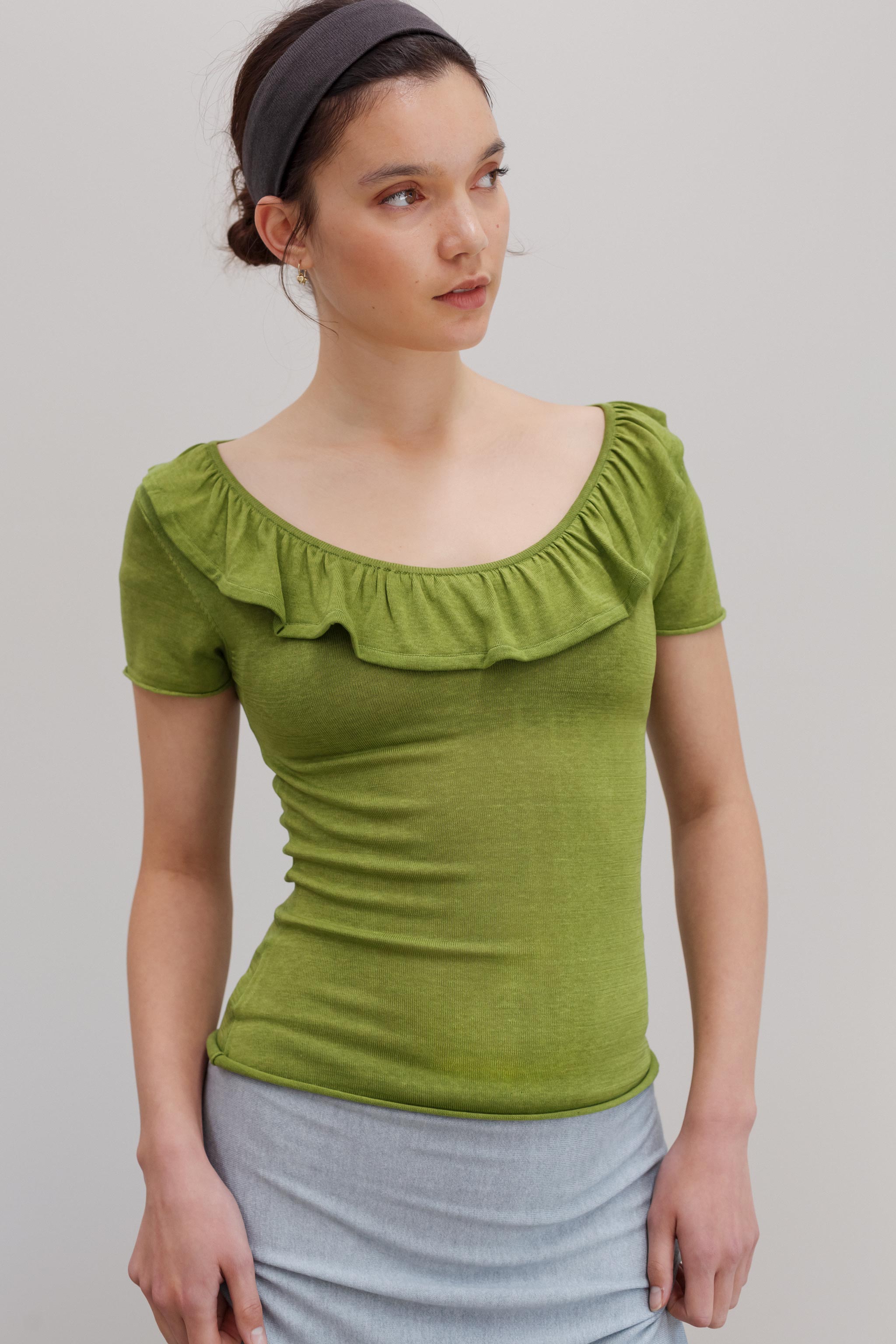 A woman wearing a fitted short-sleeve top with a wide ruffled neckline in olive green, paired with a light gray skirt, against a plain light gray studio background.