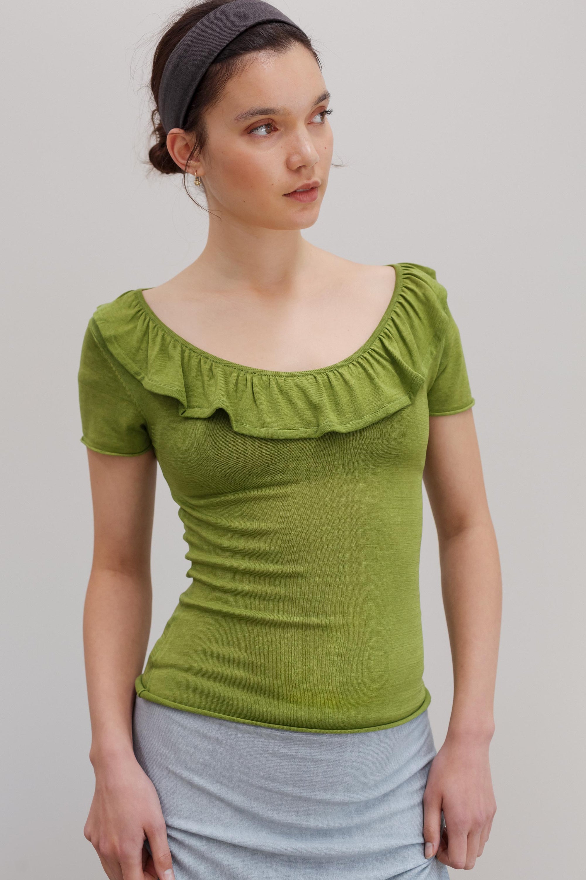 A woman wearing a fitted short-sleeve top with a wide ruffled neckline in olive green, paired with a light gray skirt, against a plain light gray studio background.
